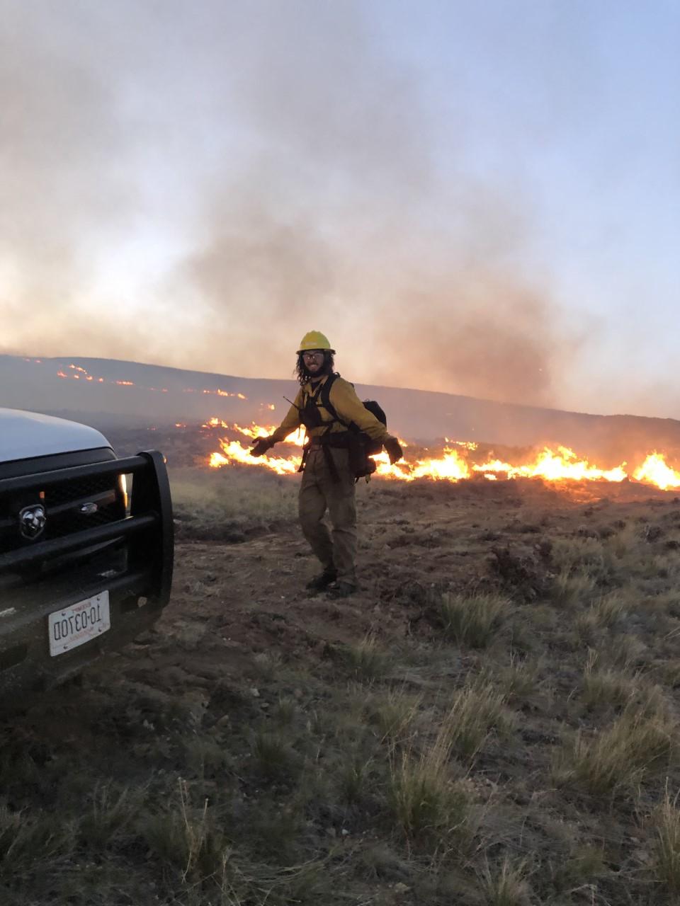 Lanning, wearing protective fire gear, smiling in a field with fire behind him. 